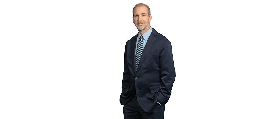 A man in a navy suit and blue tie standing with hands in pockets.