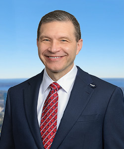 Professional portrait of a smiling man in a suit with a blue sky background.