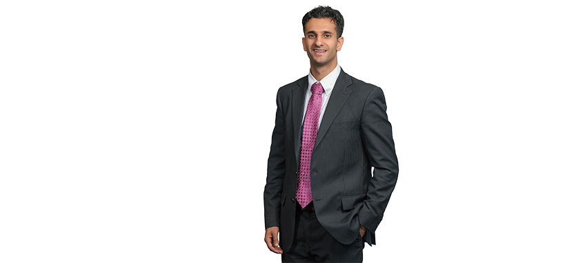 A man in a gray suit with a pink tie, smiling and standing with one hand in his pocket.