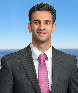 Smiling man in a suit with a pink tie outdoors against a blue sky and ocean.