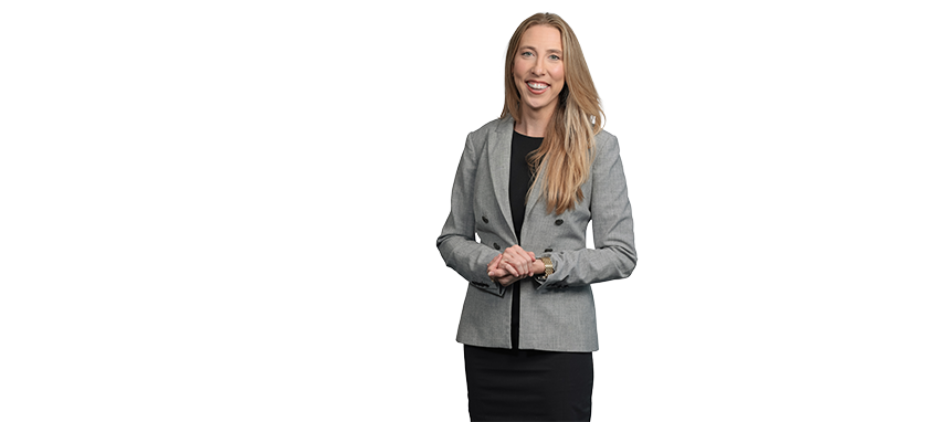 Smiling woman in gray blazer and black skirt standing with hands clasped.