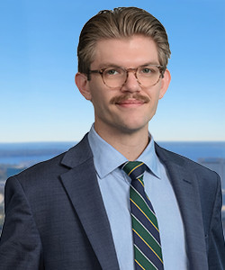Young man with glasses, mustache, in a suit and tie, outdoors with blue sky.